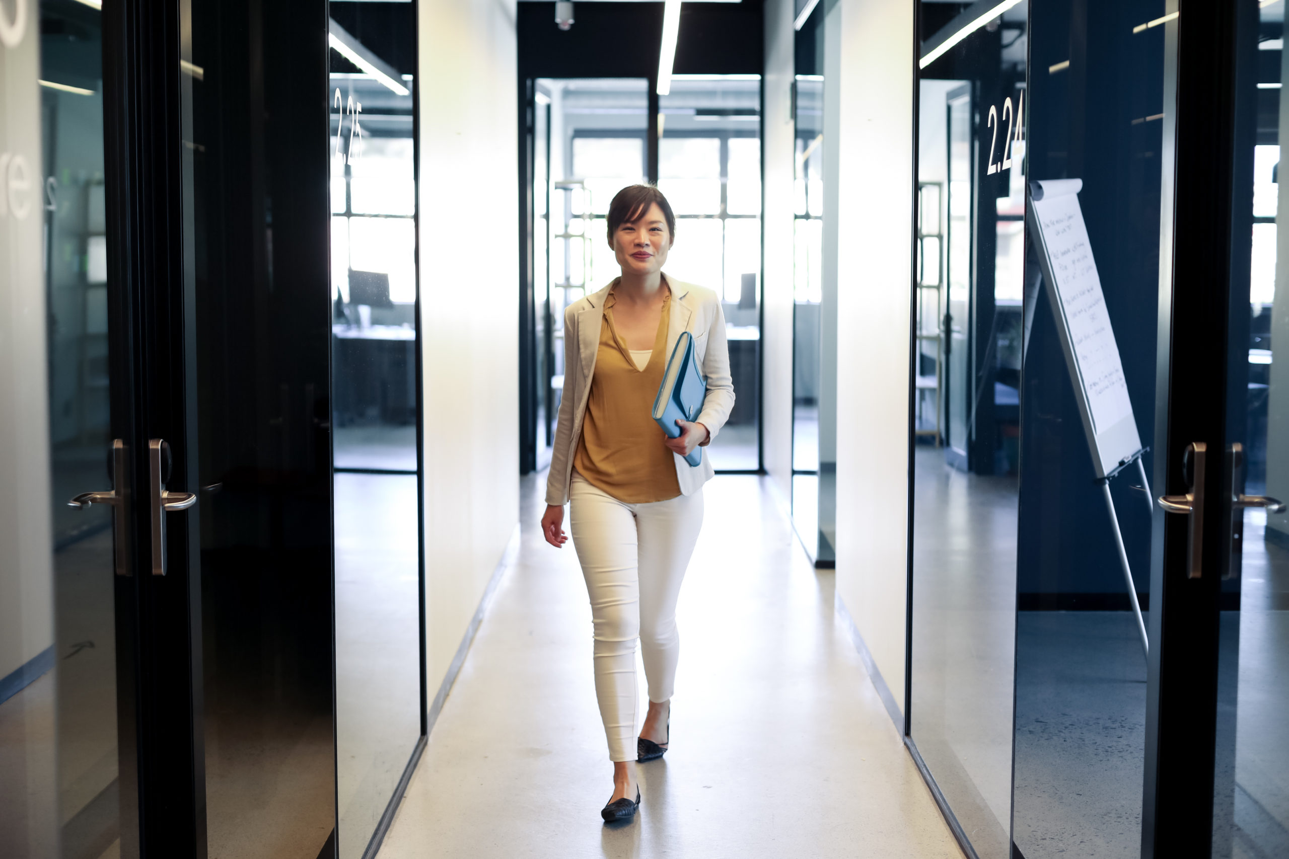 Front view of an Asian female business creative walking in the corridor of a modern office, carrying a folder and smiling to camera